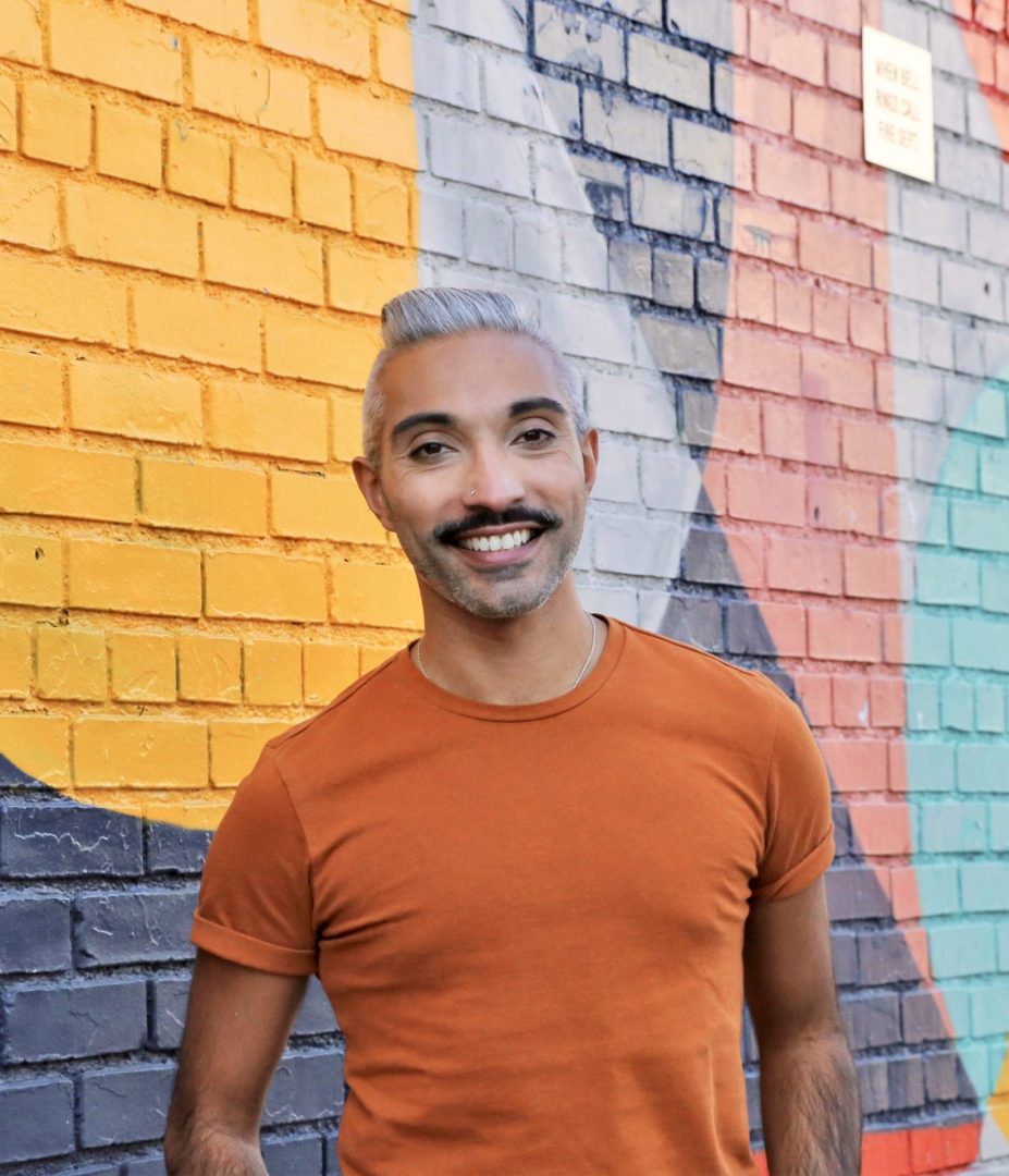 Amit standing in front of a colorful, abstract mural wearing an orange t-shirt and smiling.
